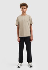 Jongen met krullend haar staat rechtop met zijn gezicht naar voren en draagt een beige t-shirt, zwarte broek en witte sneakers tegen een effen lichtgrijze achtergrond.