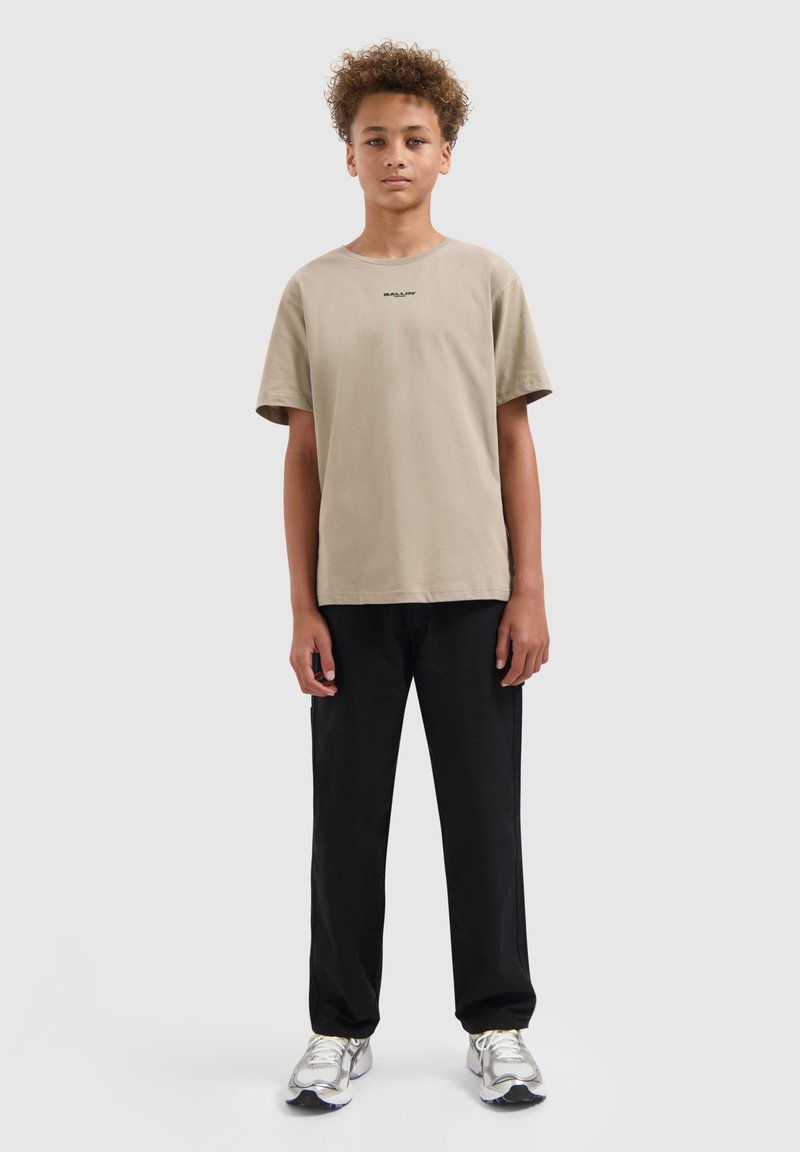 Jongen met krullend haar staat rechtop met zijn gezicht naar voren en draagt een beige t-shirt, zwarte broek en witte sneakers tegen een effen lichtgrijze achtergrond.