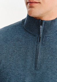 Ciemnoniebieski sweter z wysokim kołnierzem i pół-zamkiem. Tekstura materiału jest gładka, a zamek błyskawiczny ma pleciony sznurek do ciągnięcia.