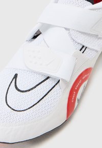 Chaussure de sport blanche avec tige en mesh, bande Velcro, détail de logo swoosh noir, accent rouge avec le texte "CYCLE SWEAT REPEAT" et semelle texturée.