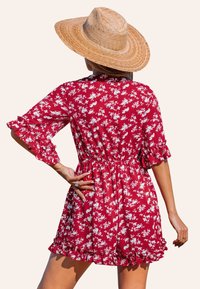 Abito rosso a fiori con maniche e orlo arricciati, vita stretta, realizzato in tessuto leggero. Indossato con un cappello di paglia a falda larga.
