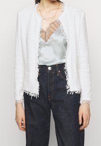 Biała teksturowana kurtka z frędzlami na krawędziach, noszona na jasnoniebieskim satynowym topie, zestawiona z ciemnymi denimowymi spodniami.