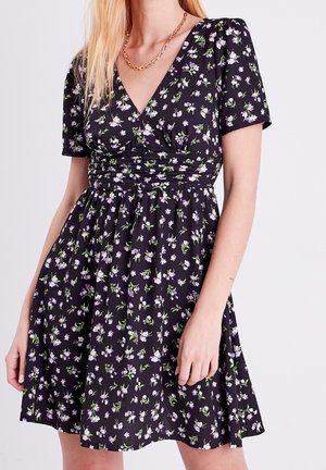 Robe noire à manches courtes avec des fleurs violettes et blanches, encolure en V, taille froncée, portée avec un collier chaîne en or.