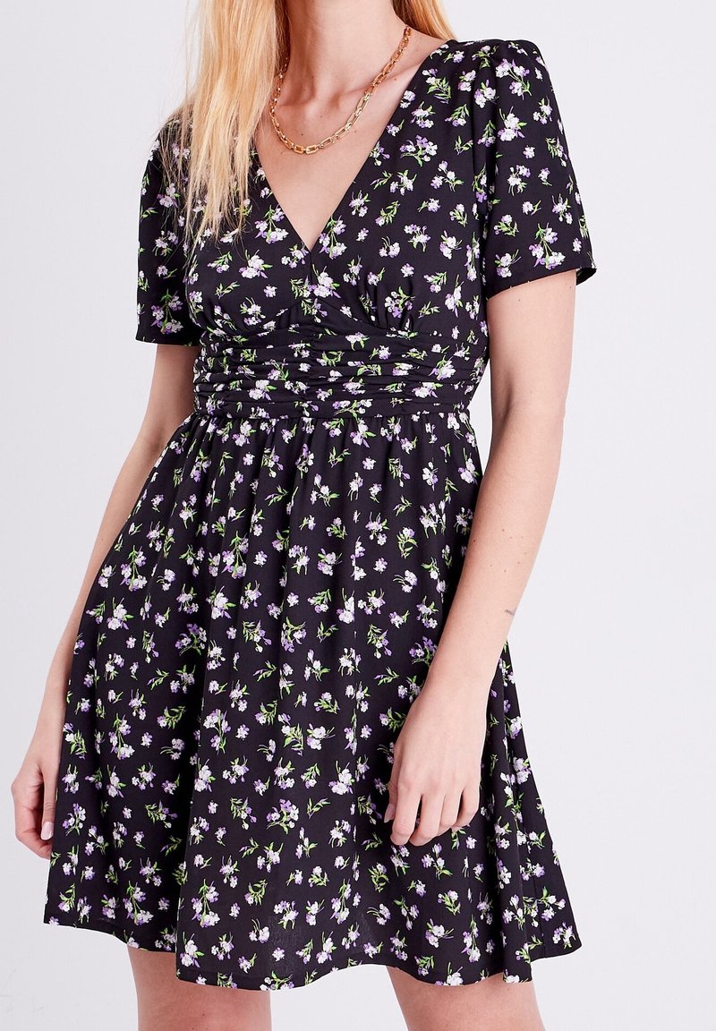 Robe noire à manches courtes avec des fleurs violettes et blanches, encolure en V, taille froncée, portée avec un collier chaîne en or.