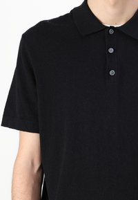 Černé polo tričko vyrobené z pletené látky. Má límečkový výstřih, krátké rukávy a zapínání na tři knoflíky se šedými knoflíky.