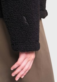Main détendue sur le côté portant une manche en polaire texturée noire et un pantalon ou une jupe marron.