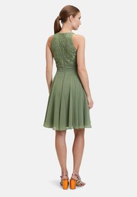 Robe verte avec un corsage en dentelle, jupe plissée et dos nu. Portée avec des sandales à talons multicolores.