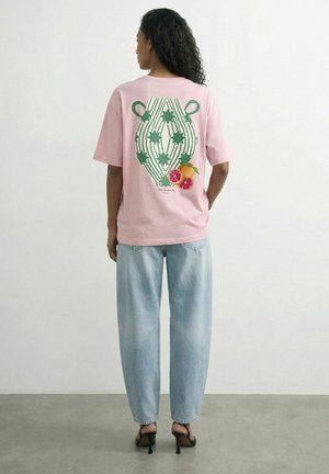 Kvinde med krøllet hår iført lyserød T-shirt med grønt stjernemønster og frugt grafik på ryggen, lyseblå jeans og sorte hæle.