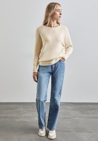 Hellbeige Strickpullover mit Rundhalsausschnitt, kombiniert mit mittelblauen Jeans im geraden Schnitt und weißen Turnschuhen. Schlichte Textur.