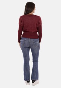 Maglione bordeaux con texture e orlo a costine, abbinato a jeans a zampa d'elefante blu. Il modello indossa scarpe da ginnastica bianche con dettagli neri.
