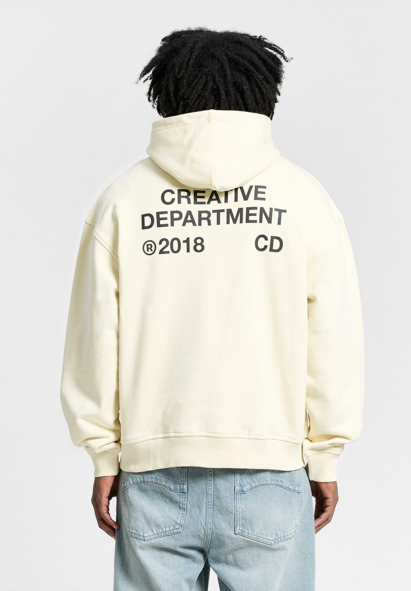 Cremefarbener Hoodie mit Kapuze und gerippten Bündchen; auf der Rückseite mit großen schwarzen Buchstaben bedruckt: "CREATIVE DEPARTMENT 2018 CD." Weicher Stoff.