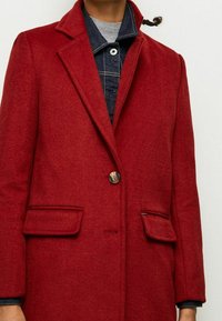 Czerwony płaszcz z mieszanki wełny z klapowanym kołnierzem, pojedynczym zapięciem na guzik i dwoma przednimi kieszeniami. Tkanina ma miękką fakturę i klasyczny fason.