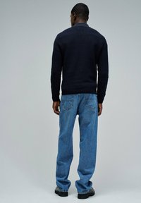 Suéter de punto azul marino, con cuello redondo, puños acanalados, combinado con jeans de corte relajado en azul claro y zapatos negros de suela gruesa, visto desde atrás.
