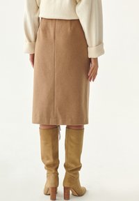 Pencil skirt in wool-blend vaaleanruskea, jossa on suora siluetti ja takasauma. Yhdistetty polven yli ulottuviin beigekuitupohjaisiin saappaisiin ja ribattuihin hihansuihin.