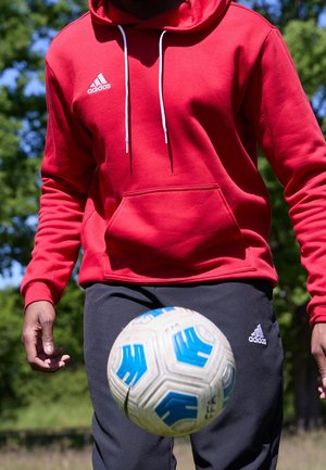 Personne portant un sweat à capuche rouge Adidas et un pantalon noir Adidas jonglant avec un ballon de football blanc et bleu en plein air, avec des arbres en arrière-plan.