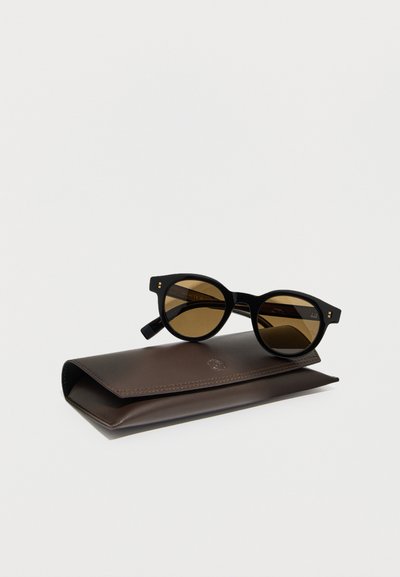 Lunettes de soleil rondes noires avec verres bruns posées sur un étui en cuir marron foncé sur un fond clair uni.