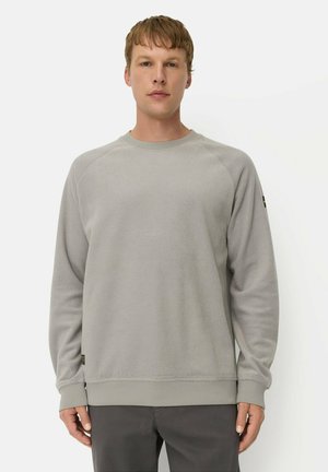 Mann mit kurzen Haaren trägt einen hellgrauen Langarm-Sweatshirt und dunkle Hosen, steht vor einem schlichten weißen Hintergrund.