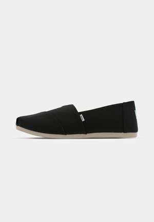 Chaussure plate noire avec une tige en toile, design de bout angulaire, brides croisées élastiquées et semelle en caoutchouc beige. Étiquette sur le côté.