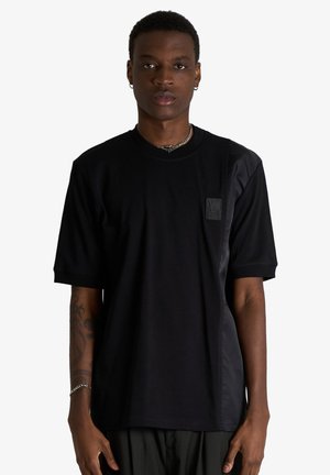 Giovane uomo che indossa una T-shirt nera a maniche corte con pannello in raso e piccolo logo sul petto, in piedi davanti a uno sfondo bianco.