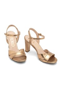 Metallisch goldene High-Heelsandalen mit offenem Zehenbereich, gekreuzten Riemen und einem Knöchelriemen mit Schnalle. Glatte Oberfläche mit einem Blockabsatzdesign.