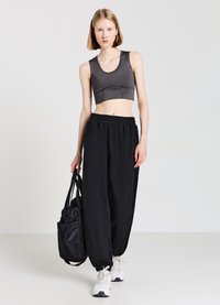 Un soutien-gorge de sport gris, court, avec un décolleté ras du cou, associé à un pantalon de jogging noir ample. Le modèle porte un sac noir et des baskets blanches.