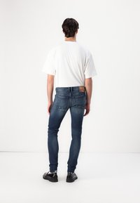 Slim fit kék farmernadrág, elhalványított mosással, öt zsebbel és bőrből készült folttal a derékszíjban. Laza fehér pólóval párosítva.