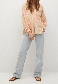 Perzikkleurige blouse met knopen en ballonmouwen, met witte bloemenpatronen; gecombineerd met lichtblauwe rechtliggende jeans en bruine sandalen.