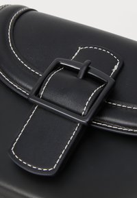 Bolso de mano de cuero negro con una solapa curva, costuras blancas y un cierre rectangular negro con una superficie texturizada.