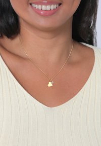 Goldenes Herz-Halskette mit einem kleinen Kristallakzent, aufgehängt an einer feinen Kette. Getragen von einer Person mit einem hellen Oberteil und glatter Haut.