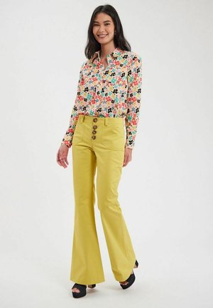 Blouse florale colorée avec un col, associée à un pantalon évasé jaune comportant trois grands boutons à l'avant. Chaussures noires à bout ouvert.