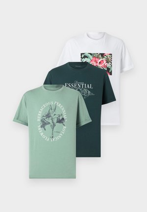Homme debout, bras croisés, portant un t-shirt vert à motif, un pantalon blanc, des chaussures noires et une montre-bracelet sur fond blanc.