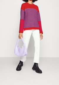 Maglione rosso lavorato a maglia con righe blu, jeans bianchi a gamba dritta, stivali neri alla caviglia e una borsa morbida color lilla. Sfondo piatto.