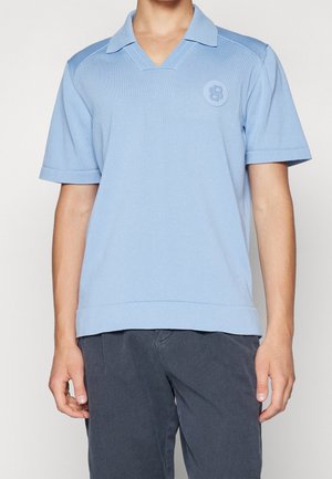 Uomo che indossa una polo a maniche corte in maglia azzurro chiaro con colletto e logo rotondo sul petto, abbinata a pantaloni grigio scuro.