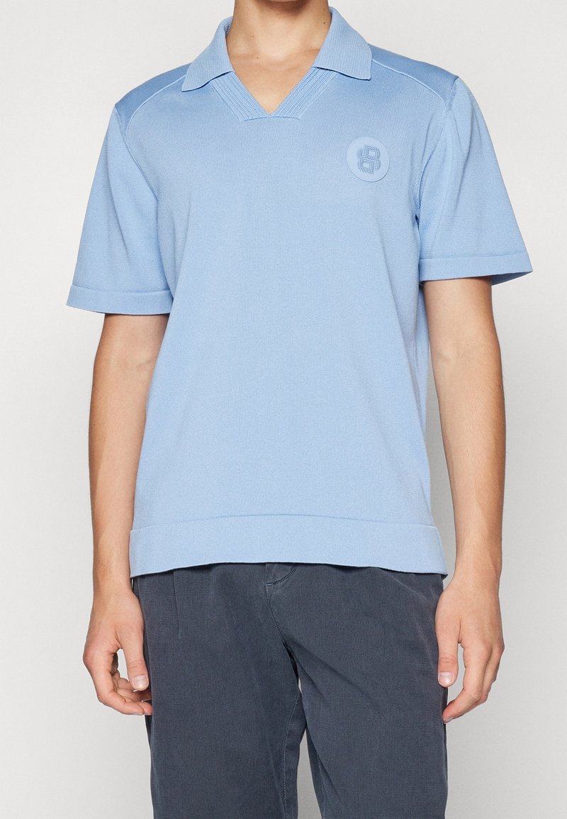 Homme portant un polo en maille bleu clair à manches courtes avec col et logo rond sur la poitrine, associé à un pantalon gris foncé.