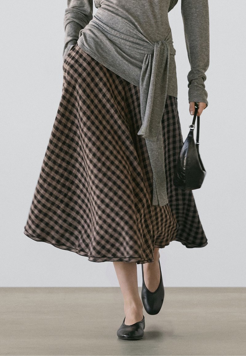 Femme portant une jupe à carreaux marron et noire, un haut gris à manches longues avec un détail noué, des ballerines noires et tenant un petit sac à main noir.