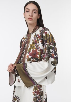 Vrouw met lang donker haar, gekleed in een witte zijden blouse met bloemenprint en een bijpassende rok met wijde mouwen en groene, rode en paarse accenten.
