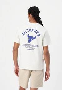 Biely tričko s modrou grafikou, na ktorej je lebka býka a text "SALTON SEA SURF CLUB" na chrbte. Krátke rukávy, voľný strih.