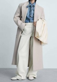 Manteau en laine beige porté sur une chemise en jean avec une cravate, accompagné d'un pantalon large crème et de chaussures claires. Le fond est gris clair.
