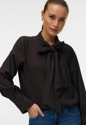 Femme portant un chemisier noir avec un grand nœud noué au cou, manches longues et jean bleu, main dans la poche.