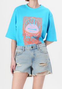 Světle modré oversized tričko s živým grafickým designem v růžové a oranžové barvě. Džínové šortky s obnošenými okraji.