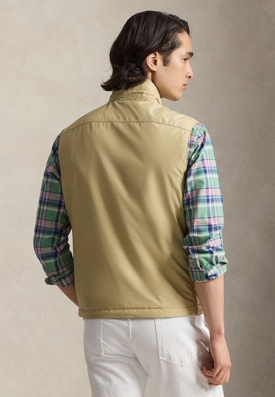 Αμάνικο μπεζ γιλέκο πάνω από ένα πράσινο και μπλε καρό πουκάμισο, με μανίκια που είναι γυρισμένα και λεία υφή. Άσπρο παντελόνι φαίνεται στο κάτω μέρος.