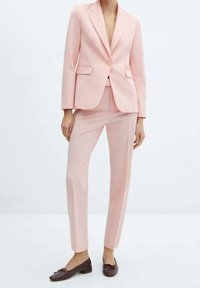 Costume sur mesure rose clair avec un blazer cintré et un pantalon slim, doté d'une fermeture à un seul bouton et de poches ; porté avec des ballerines marron.