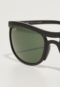 Ochelari de soare din plastic negru cu lentile verzi, având un design elegant, formă în jurul feței și o marcă subtilă pe partea de sus a ramei.