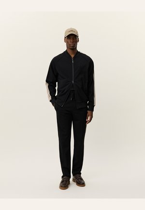 Jachetă zip-up neagră cu dungi laterale crem, asortată cu pantaloni negri. Purtați cu o șapcă bej și pantofi închiși la culoare cu tălpi de culoare maro deschis.