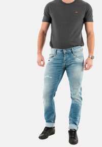 T-shirt gris pour homme à manches courtes avec un logo rouge, associé à un jean bleu clair déchiré et des baskets noires. Le jean a des ourlets retroussés.