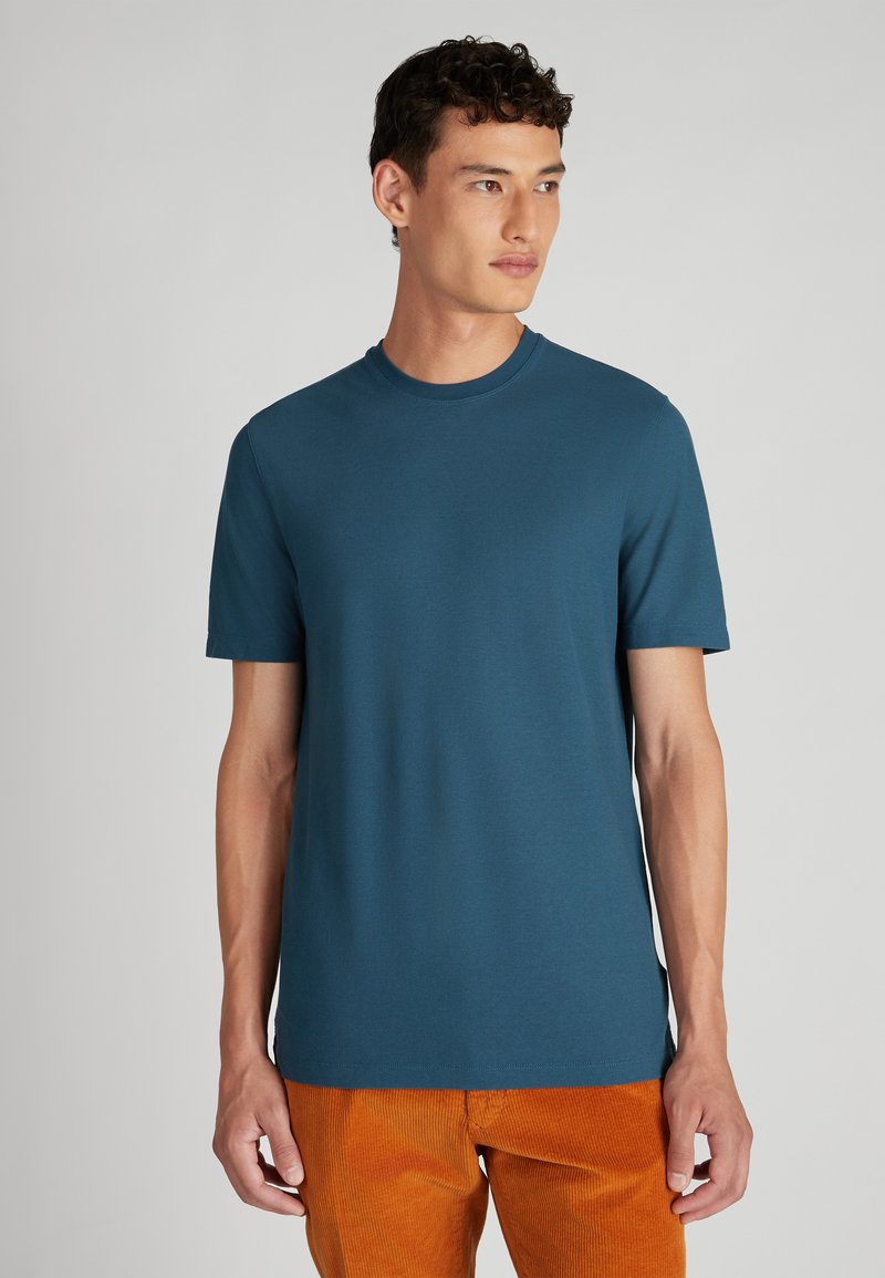 Joven con cabello rizado oscuro, vistiendo una camiseta de manga corta color teal y pantalones de pana naranjas, mirando ligeramente a la derecha.