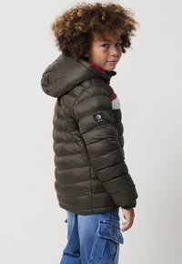 Niño con cabello rizado que lleva una chaqueta acolchada verde oscuro con capucha y pantalones cargo azul mirando por encima del hombro.