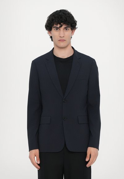 Navy blazer with structured lapels, two black buttons, and side pockets, worn over a black shirt, featuring a tailored fit.
