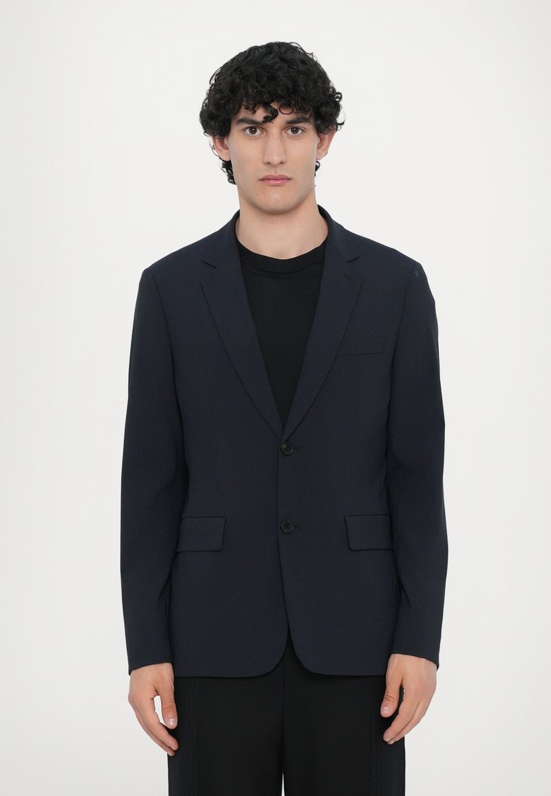 Mørkeblå blazer lavet af glat stof, med en to-knap front, notch revers og to forlommer. Båret over en sort skjorte.