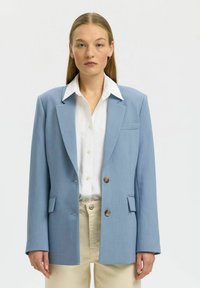 Femme aux cheveux blonds raides portant un blazer oversize bleu clair, une chemise blanche boutonnée et un pantalon beige sur un fond uni.